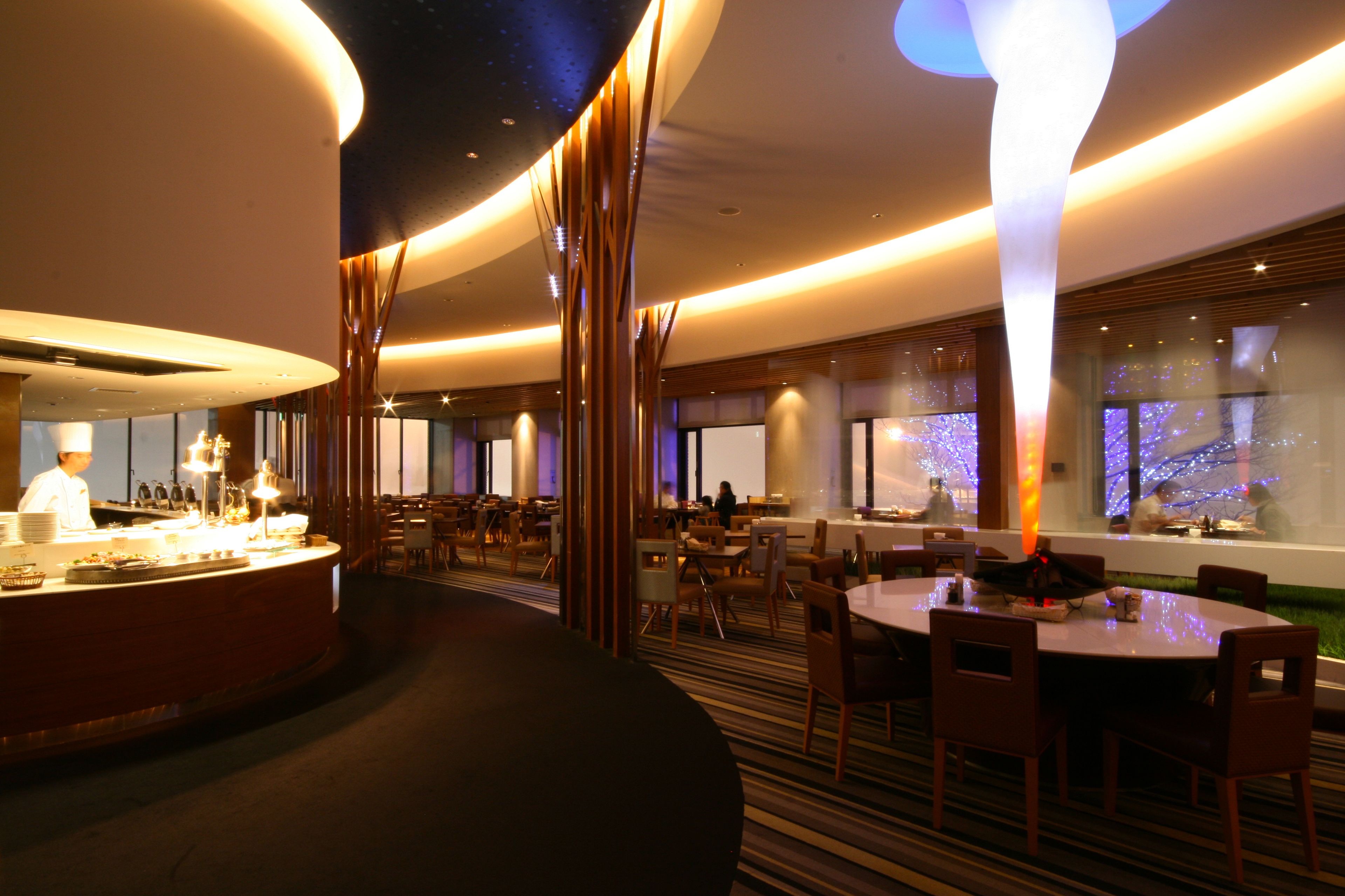 A modern restaurant with a central bar, round tables, and a chef preparing food.