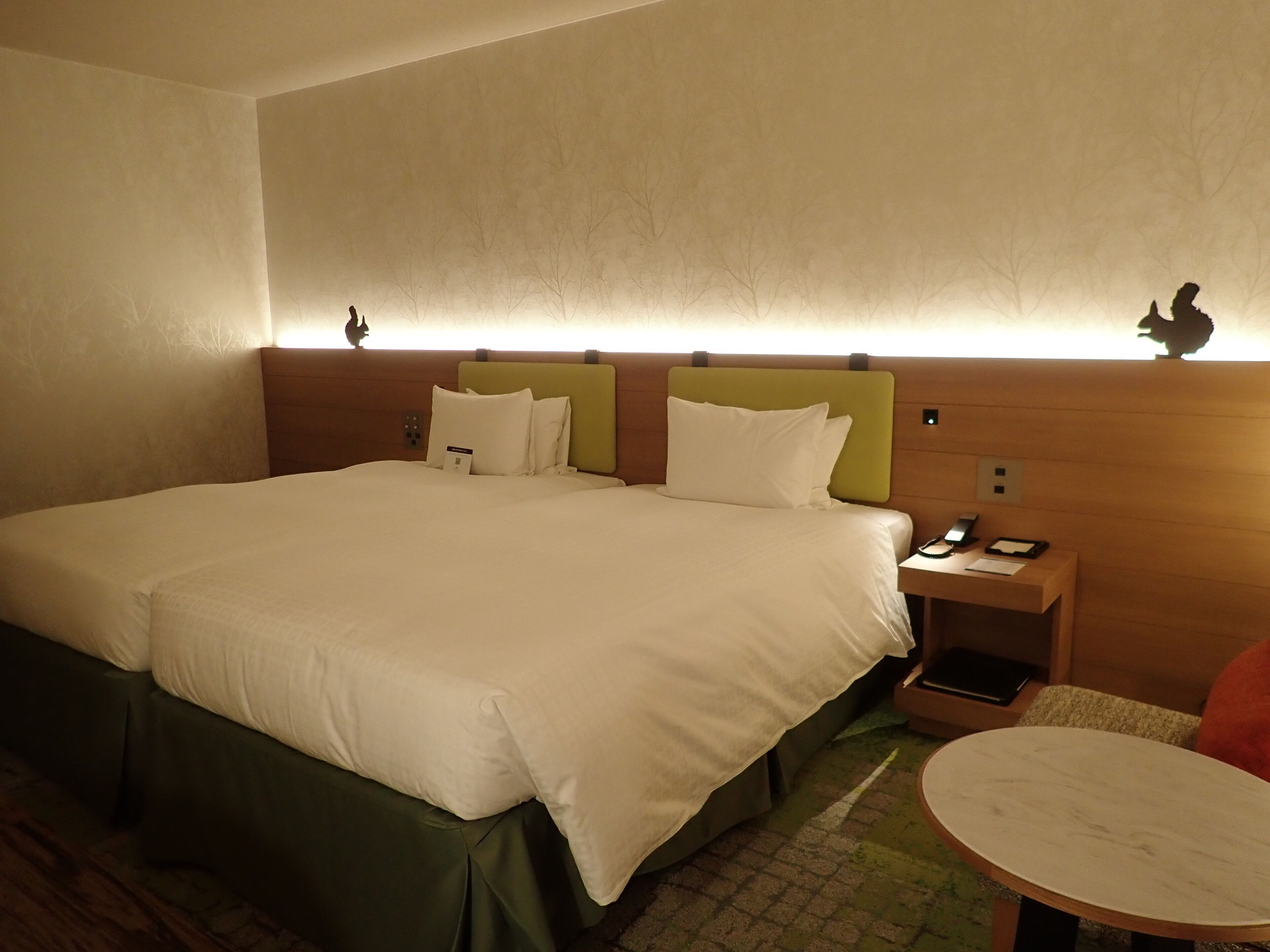 A hotel room with a large bed, a bedside table with a phone and a lamp, and a small round table with a chair.