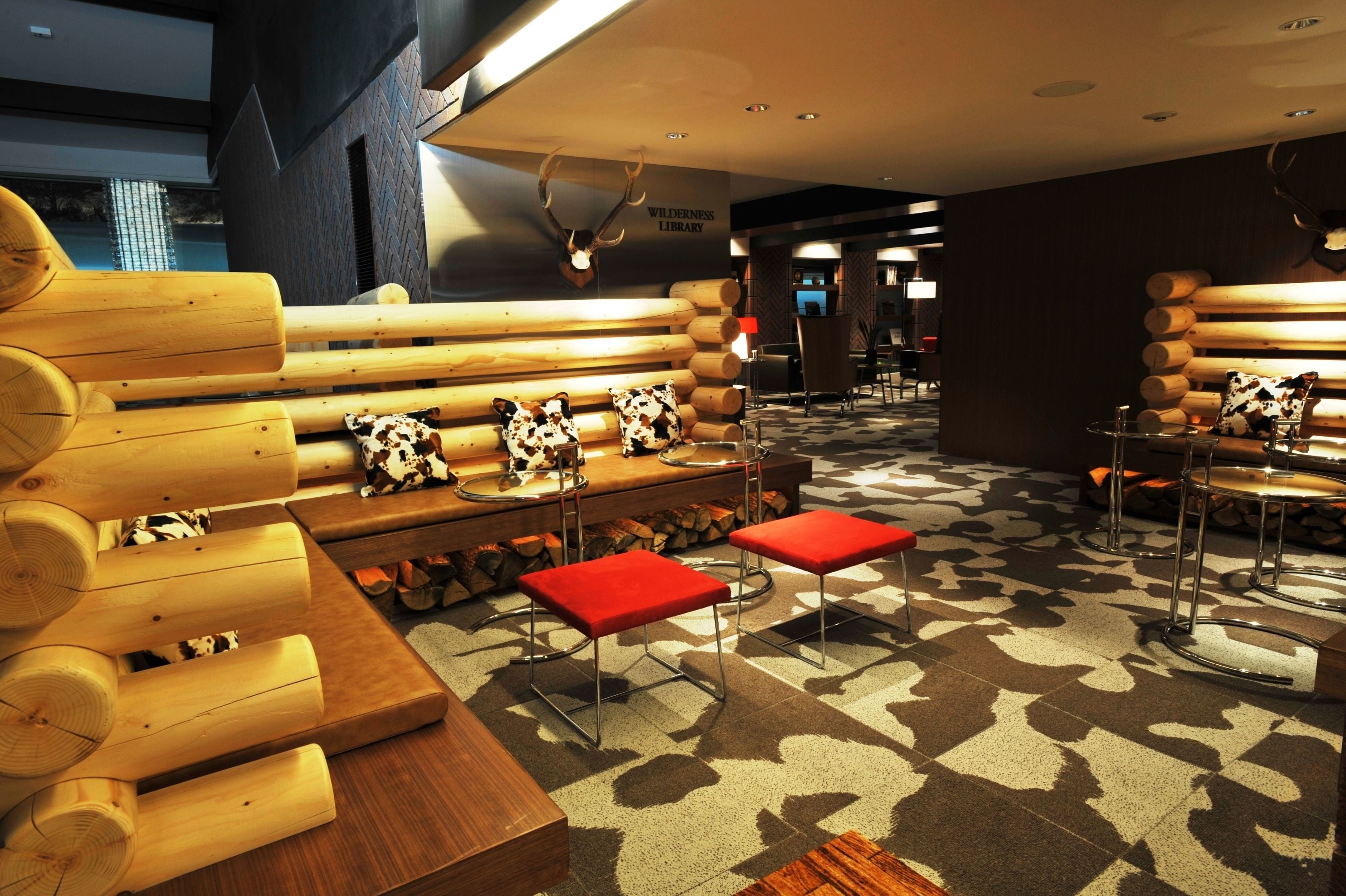 A log cabin-style interior with wooden benches and red stools, a patterned carpet, and antler decorations.