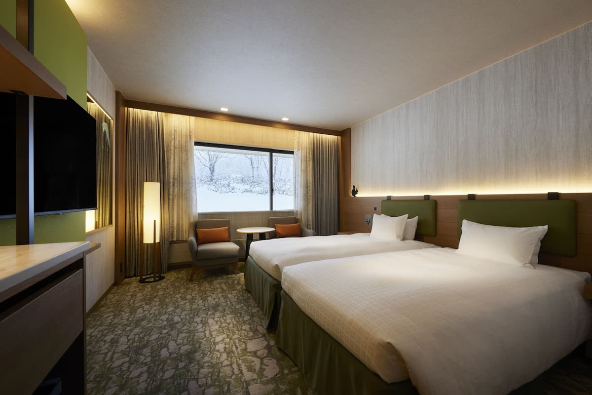 A hotel room with two beds, a desk, a chair, a small table, and a window with curtains.
