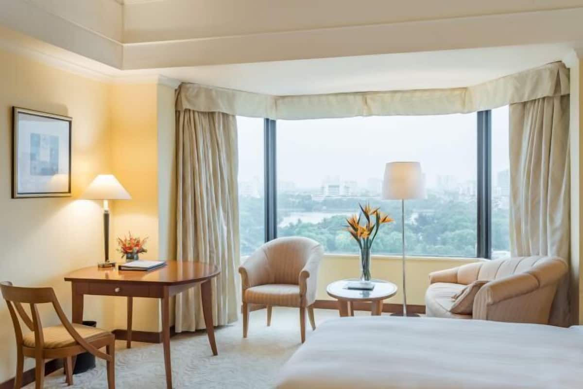 A hotel room with a bed, a desk, two chairs, a small table with a lamp, and a view of the city through large windows.
