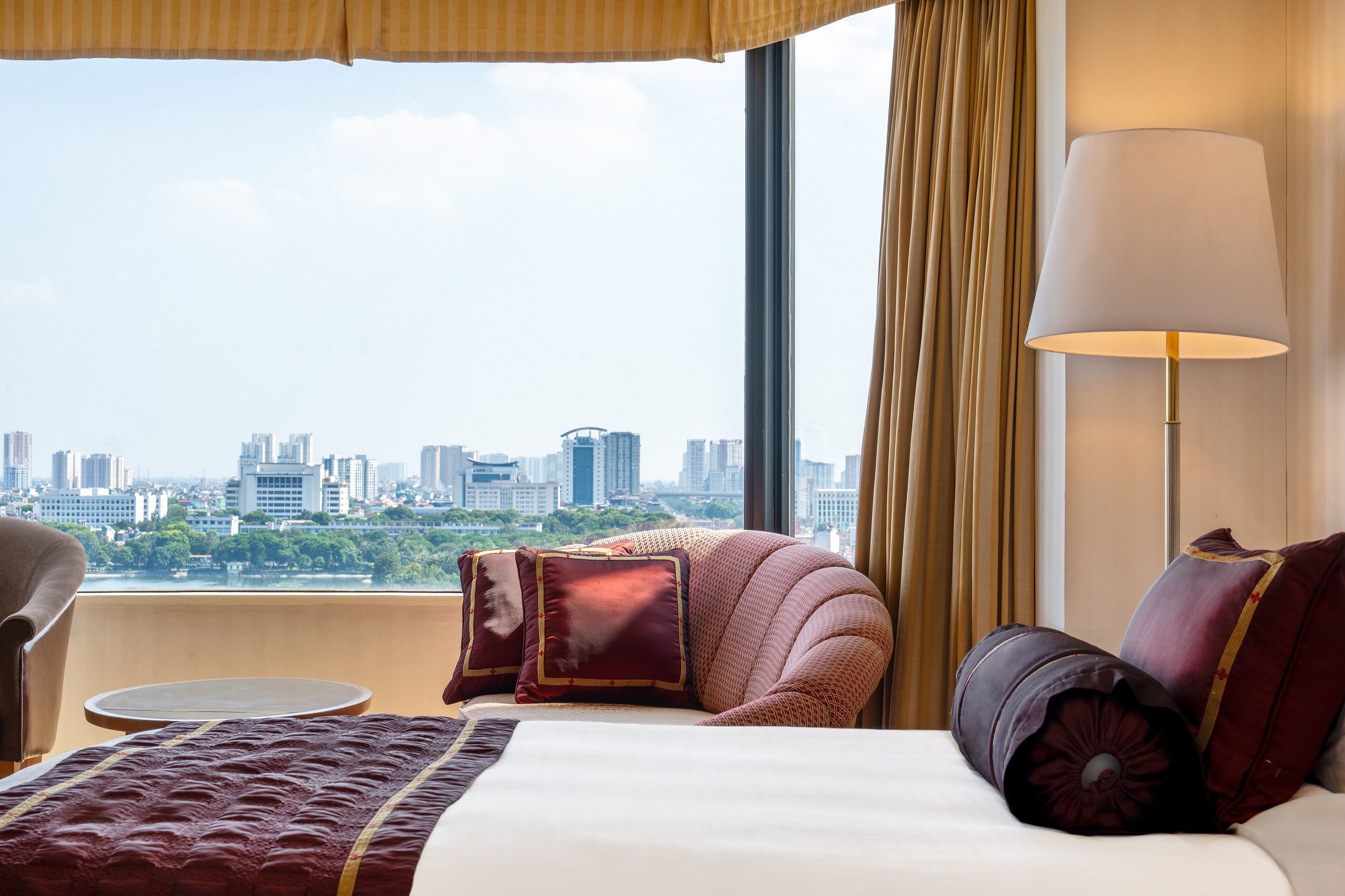 A hotel room with a large bed, a round table, a sofa, and a view of a cityscape through a large window.