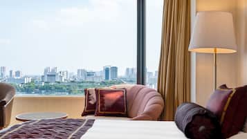 A hotel room with a large bed, a round table, a sofa, and a view of a cityscape through a large window.
