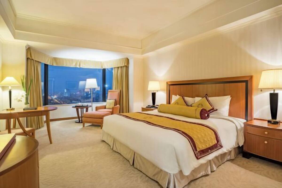 A hotel room with a large bed, two bedside tables, a desk, a chair, and a large window with a city view.