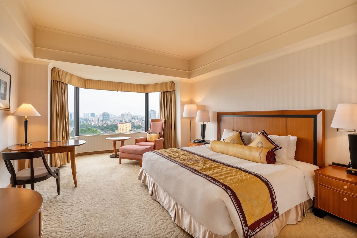 A hotel room with a large bed, a desk, a chair, and a view of a cityscape through a large window.