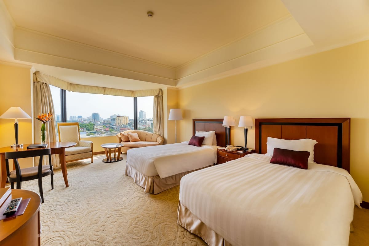 A hotel room with two beds, a desk, a chair, and a view of the city.