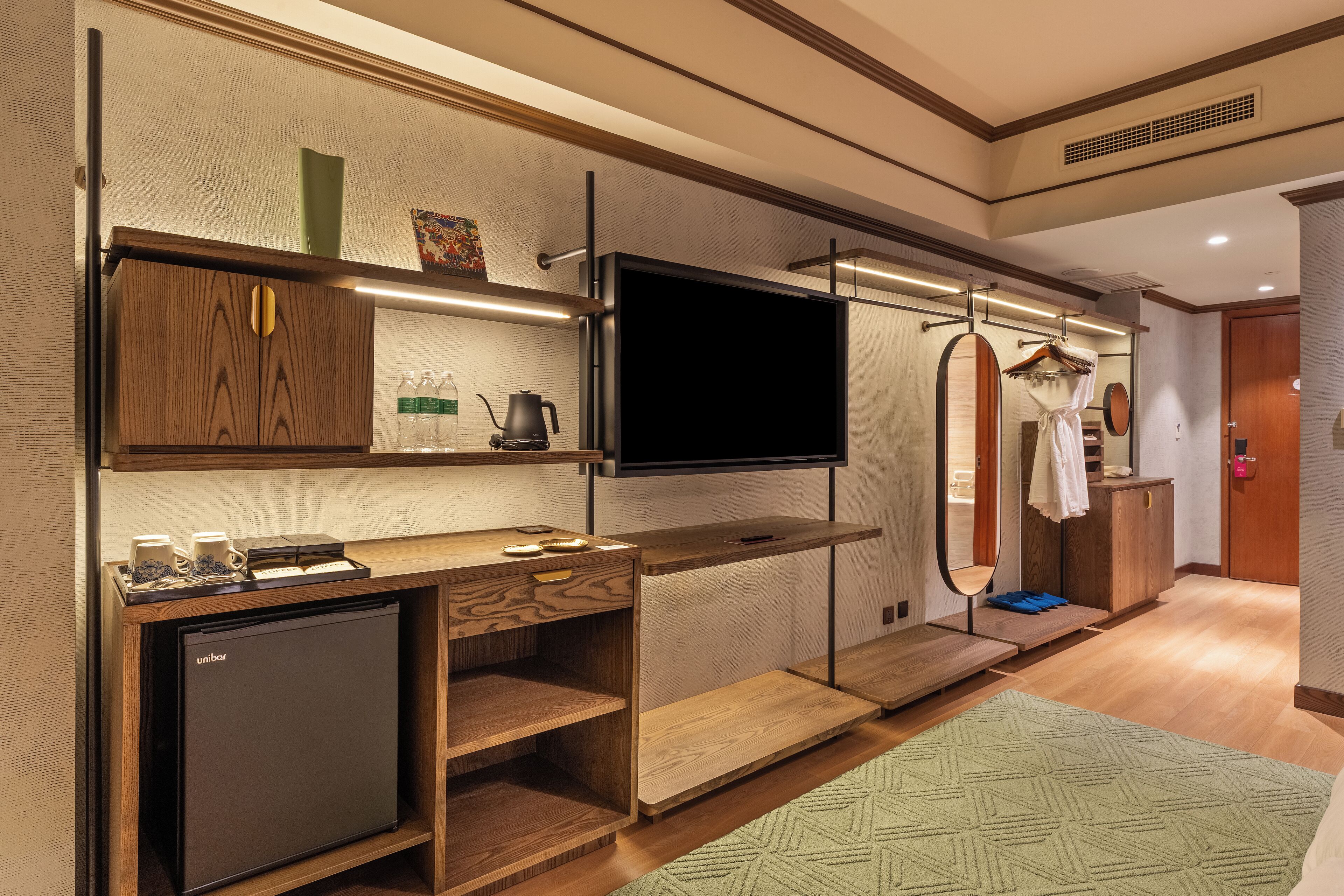 A compact hotel room with a minibar, a flat-screen TV, a wooden cabinet, and a clothing rack.