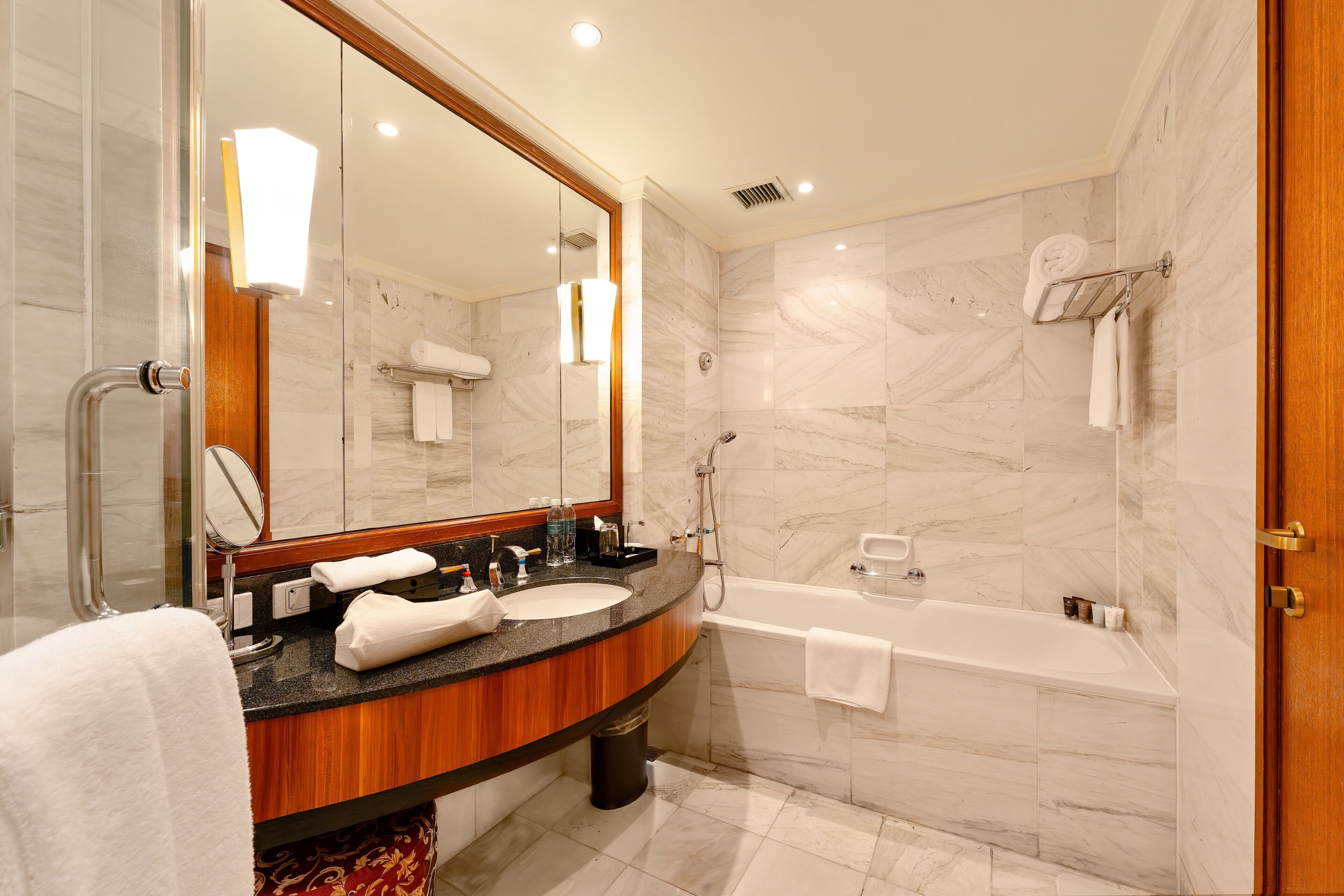 A modern bathroom with a marble wall, a wooden vanity, a large mirror, a bathtub, and a shower area.