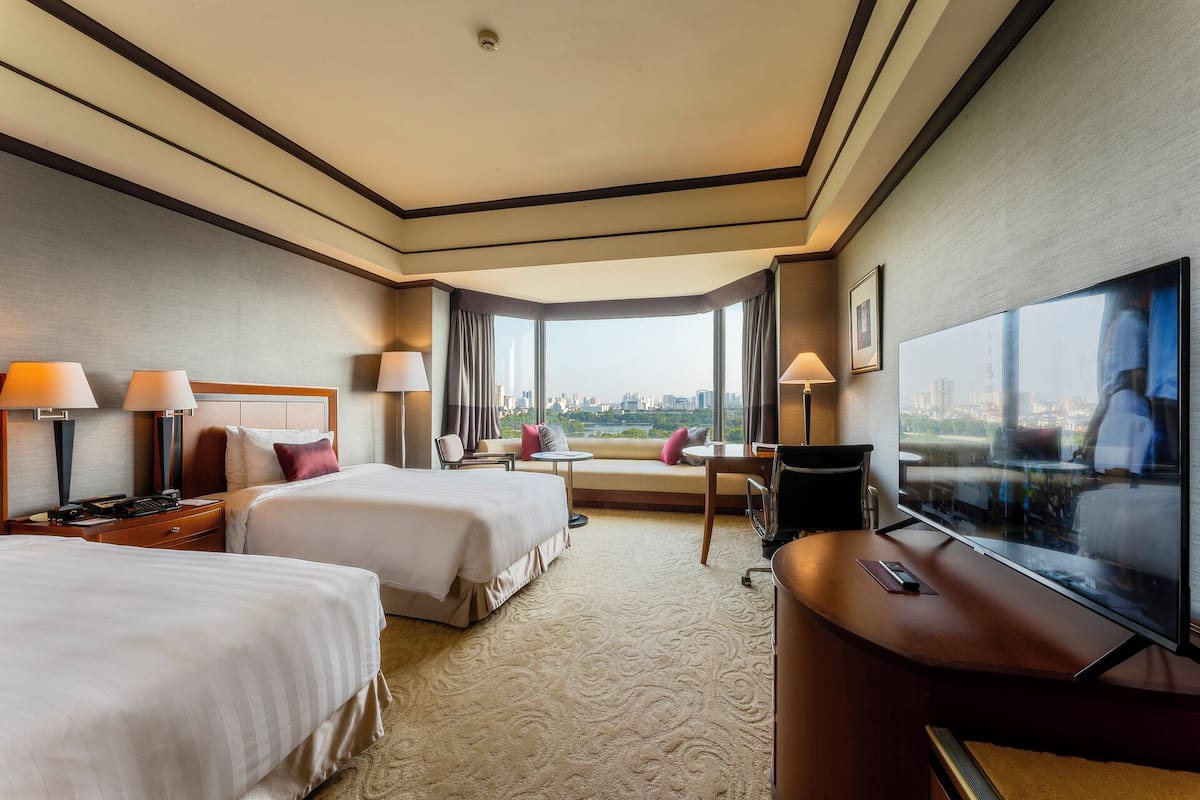 A hotel room with two beds, a desk with a TV, and a large window offering a city view.