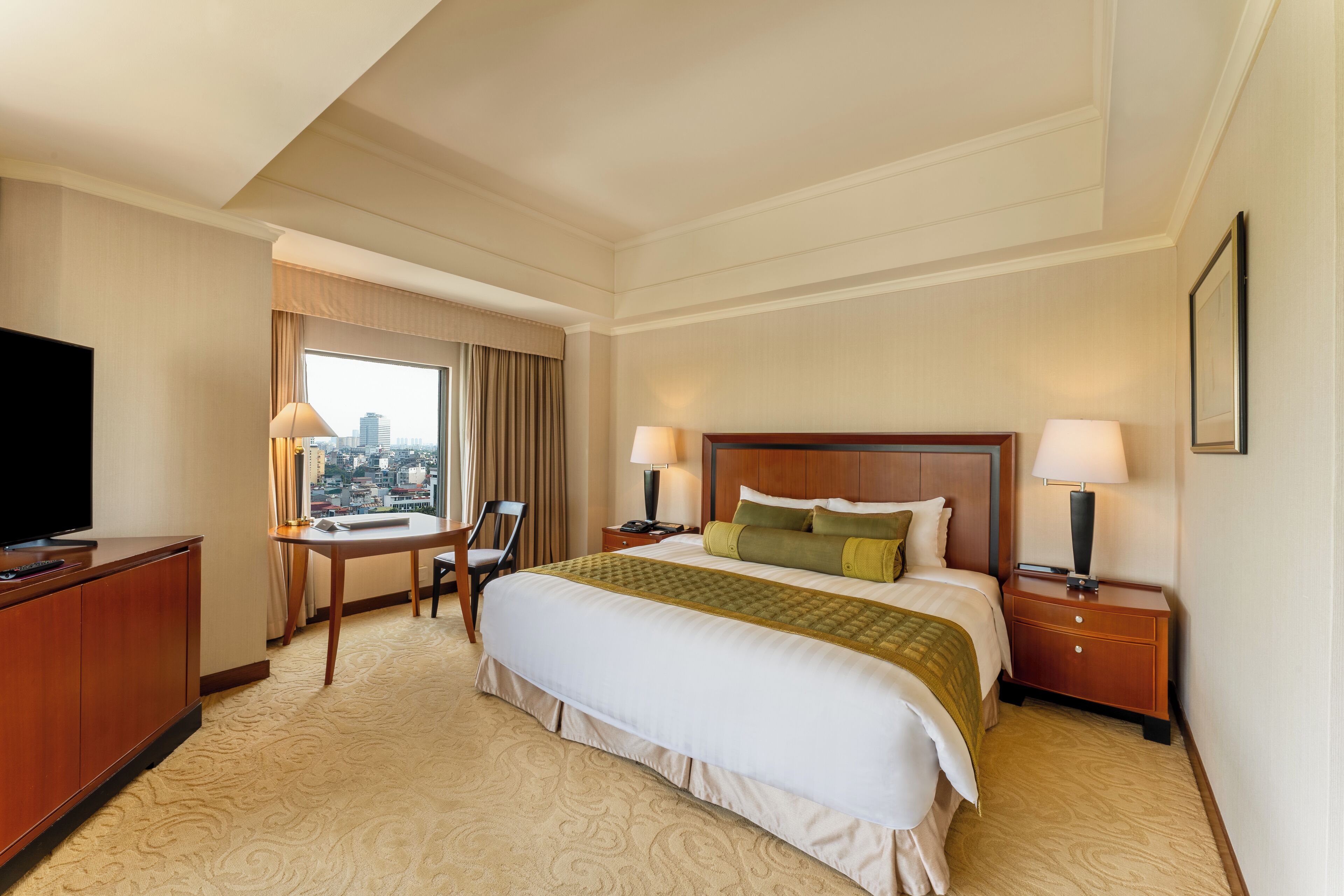 A hotel room with a large bed, two bedside tables with lamps, a desk with a chair, a television, and a window with a city view.