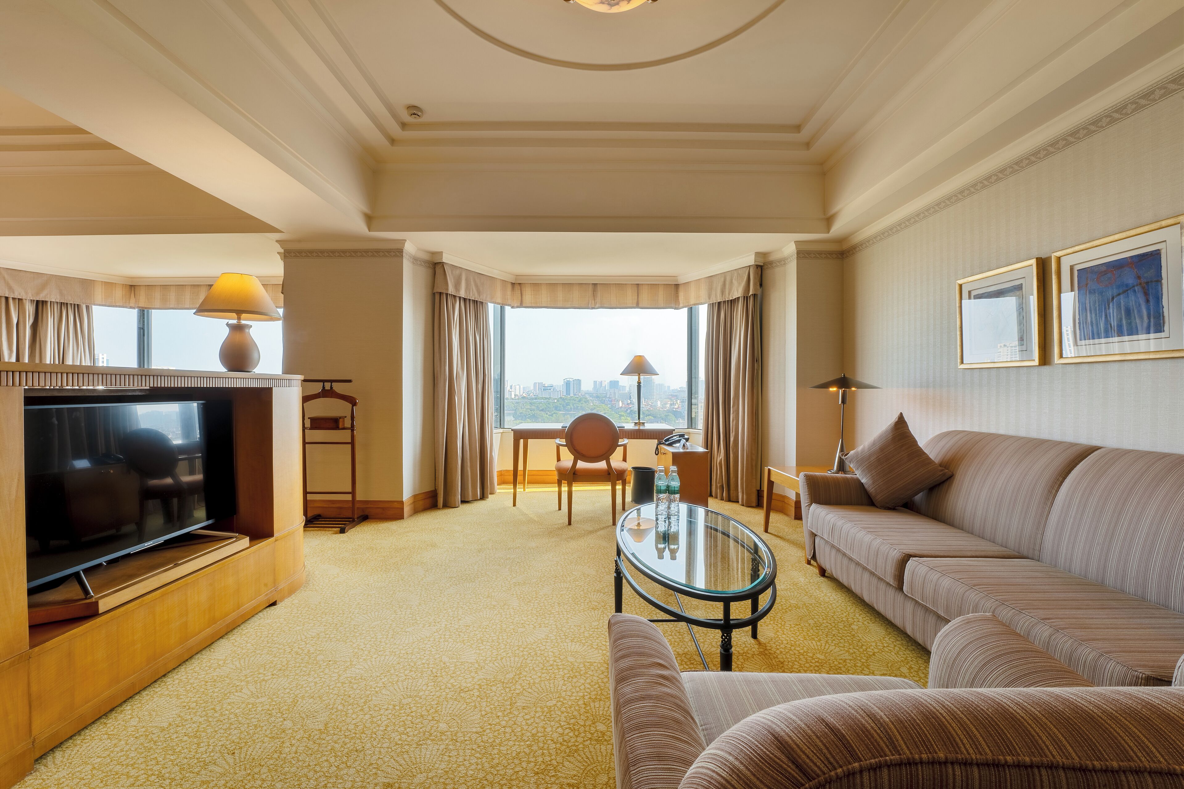 A hotel room with a TV, sofa, armchair, and a desk with a lamp.