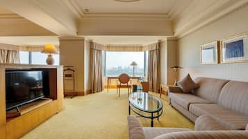 A hotel room with a TV, sofa, armchair, and a desk with a lamp.