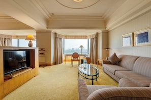A hotel room with a TV, sofa, armchair, and a desk with a lamp.
