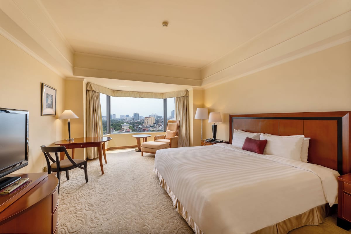 A hotel room with a large bed, a desk with a TV, a chair, a small table, and a view of the city through a large window.