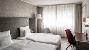 A hotel room with two beds, a desk, a chair, a lamp, and a large window with curtains.