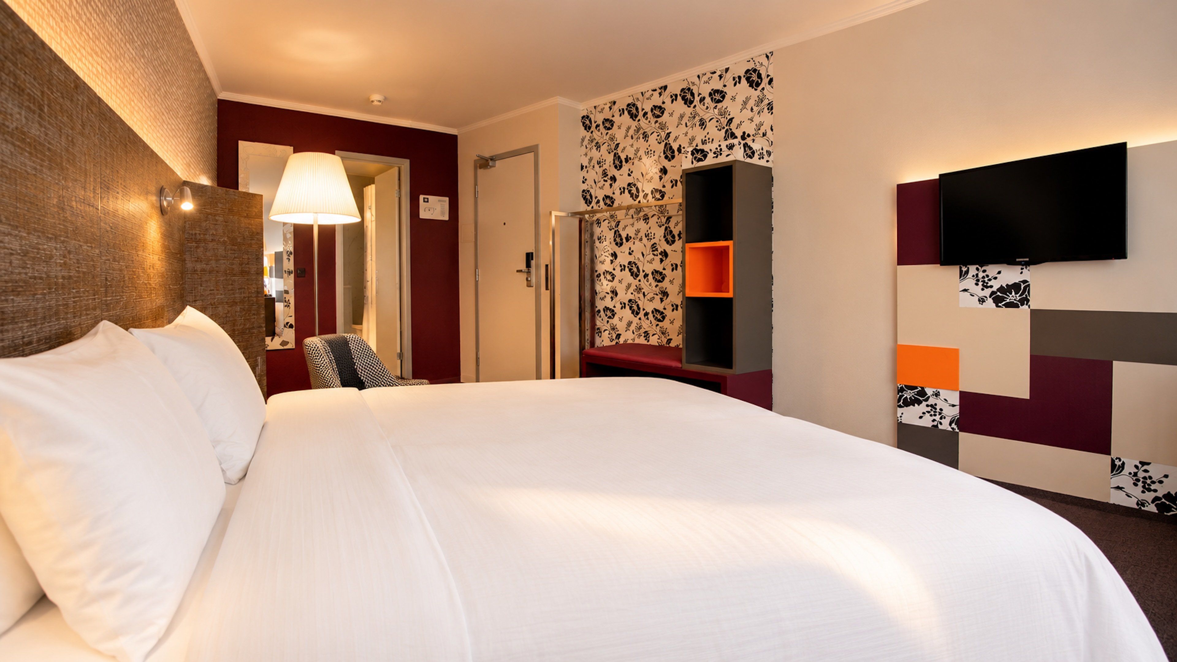 Habitación de hotel con una cama grande, un televisor de pantalla plana montado en la pared, un escritorio con una silla y una pequeña zona de estar con un banco.