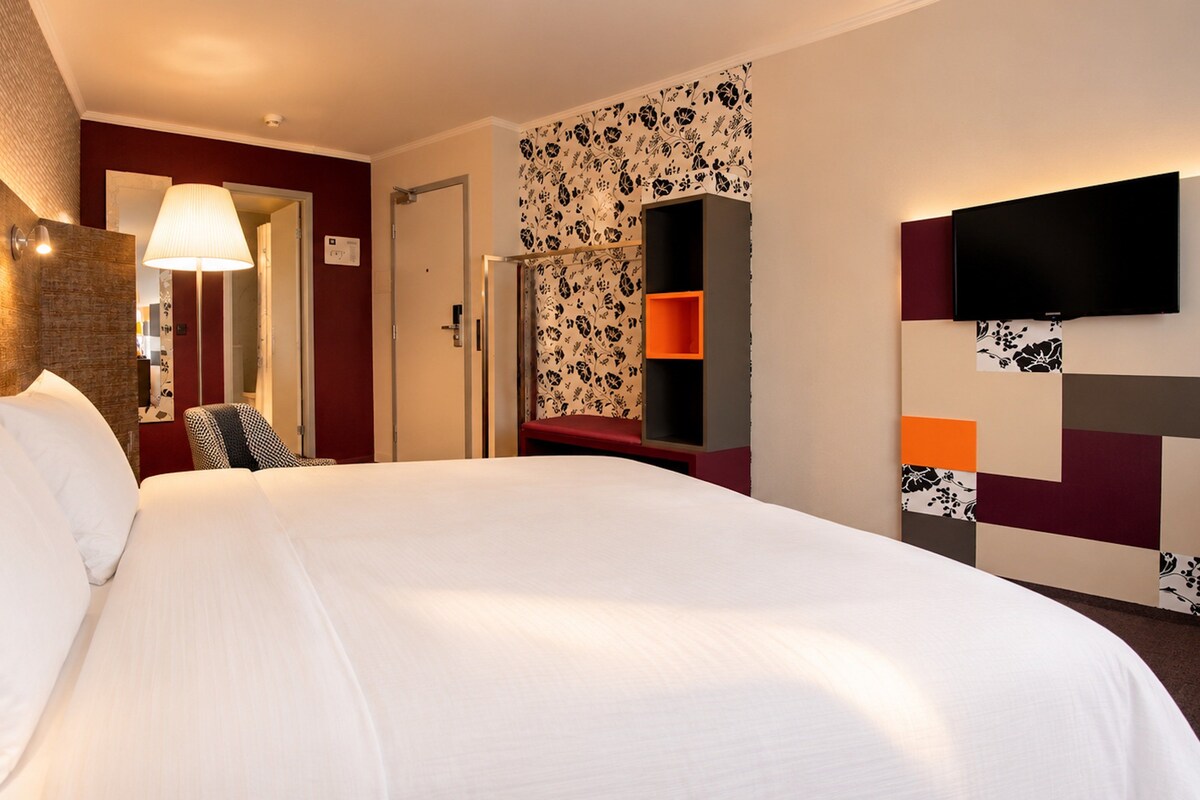 A hotel room with a large bed, a flat-screen TV mounted on the wall, a desk with a chair, and a small seating area with a bench.
