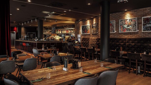 A restaurant with wooden tables, grey chairs, a bar area with shelves of bottles, and a brick wall with framed pictures. - pentahotel Kassel (Kassel)