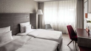 A hotel room with two beds, a desk, a chair, a lamp, and a large window with curtains. - pentahotel Kassel (Kassel)
