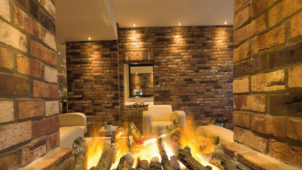 A cozy room with a brick wall, a fireplace with logs, and two white chairs.