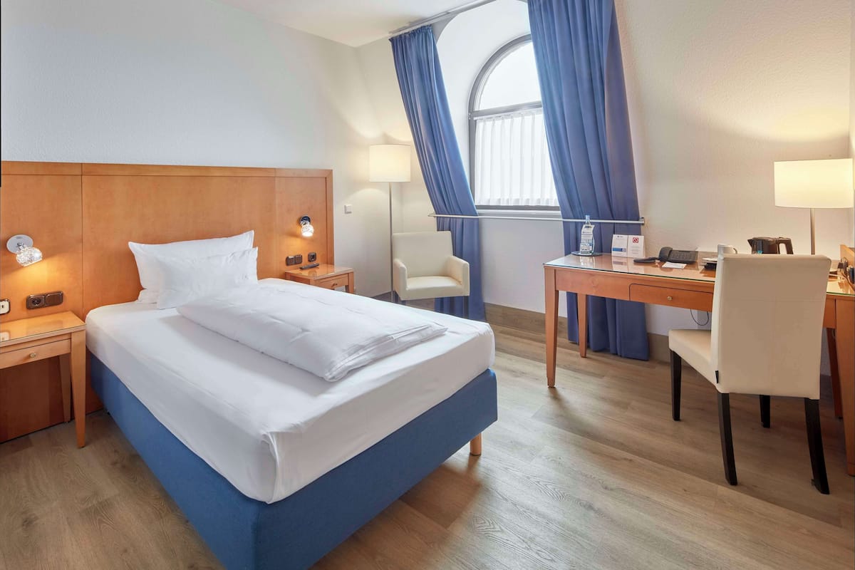 A hotel room with a bed, bedside tables, a desk, a chair, and a window with curtains.