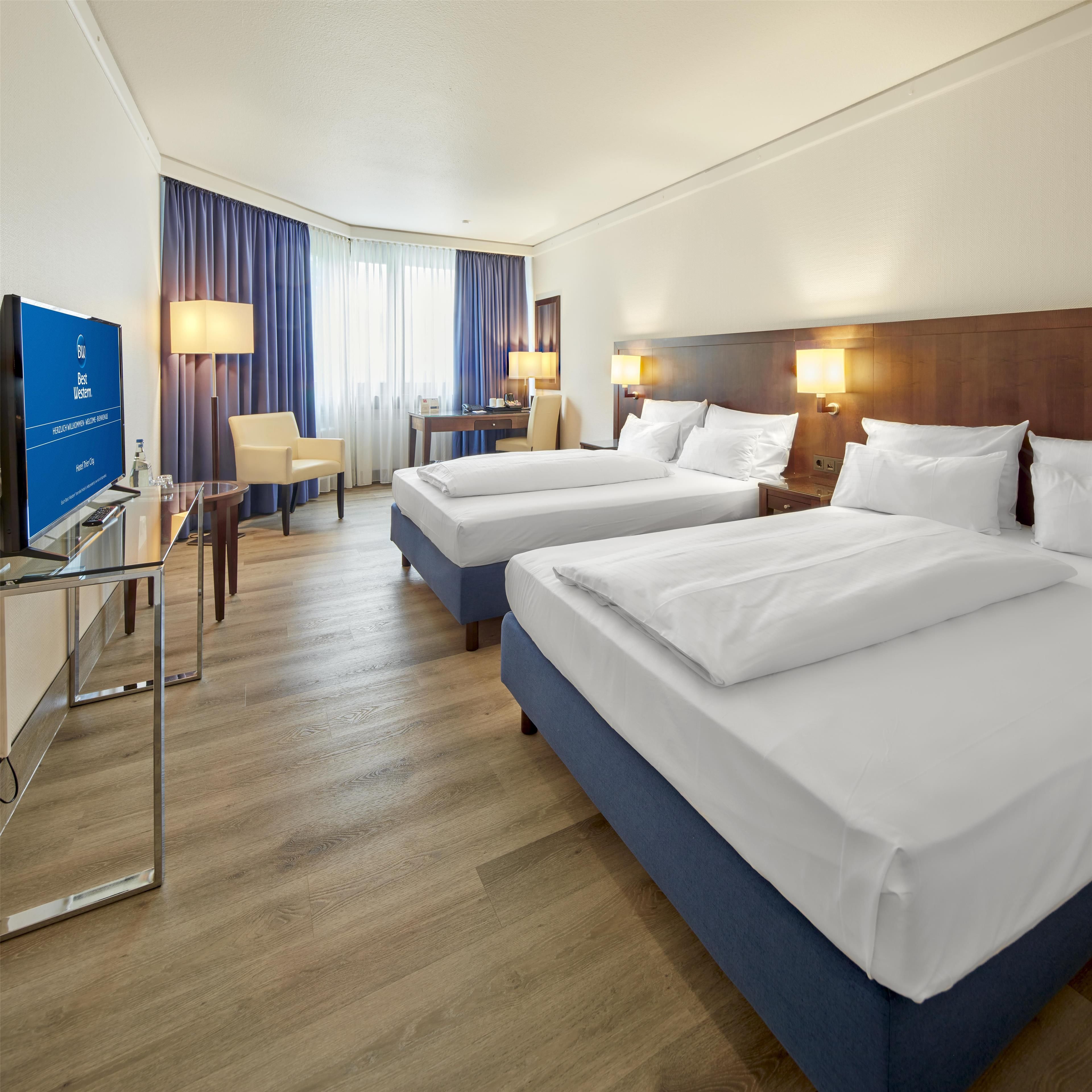 Ein Hotelzimmer mit zwei Betten, einem Schreibtisch, einem Sessel, einem Fernseher und einem Fenster mit Vorhängen.