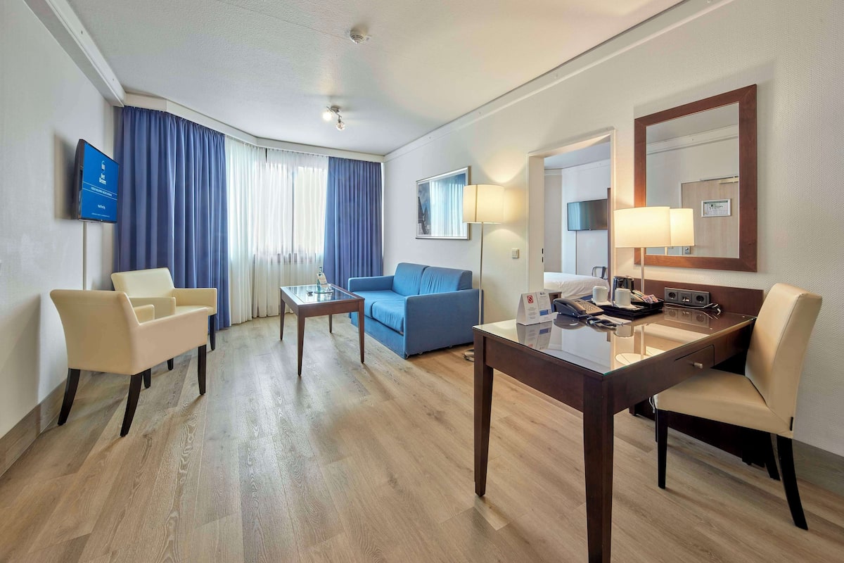 A hotel room with a wooden floor, a blue sofa, a beige armchair, a glass coffee table, a desk with a telephone, and a flat-screen TV mounted on the wall.