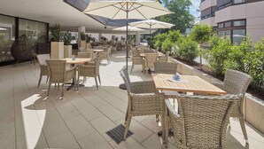 An outdoor patio area with wicker furniture and large umbrellas, surrounded by greenery and modern buildings.