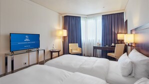 Une chambre d’hôtel avec deux grands lits, un bureau, deux chaises, un téléviseur affichant un écran d’accueil et une fenêtre avec des rideaux.