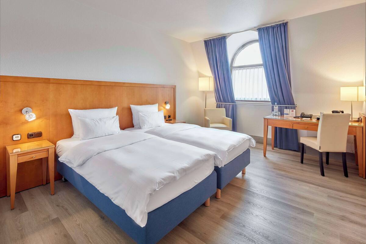 A hotel room with a double bed, a desk with a chair, a lamp, and a window with curtains.