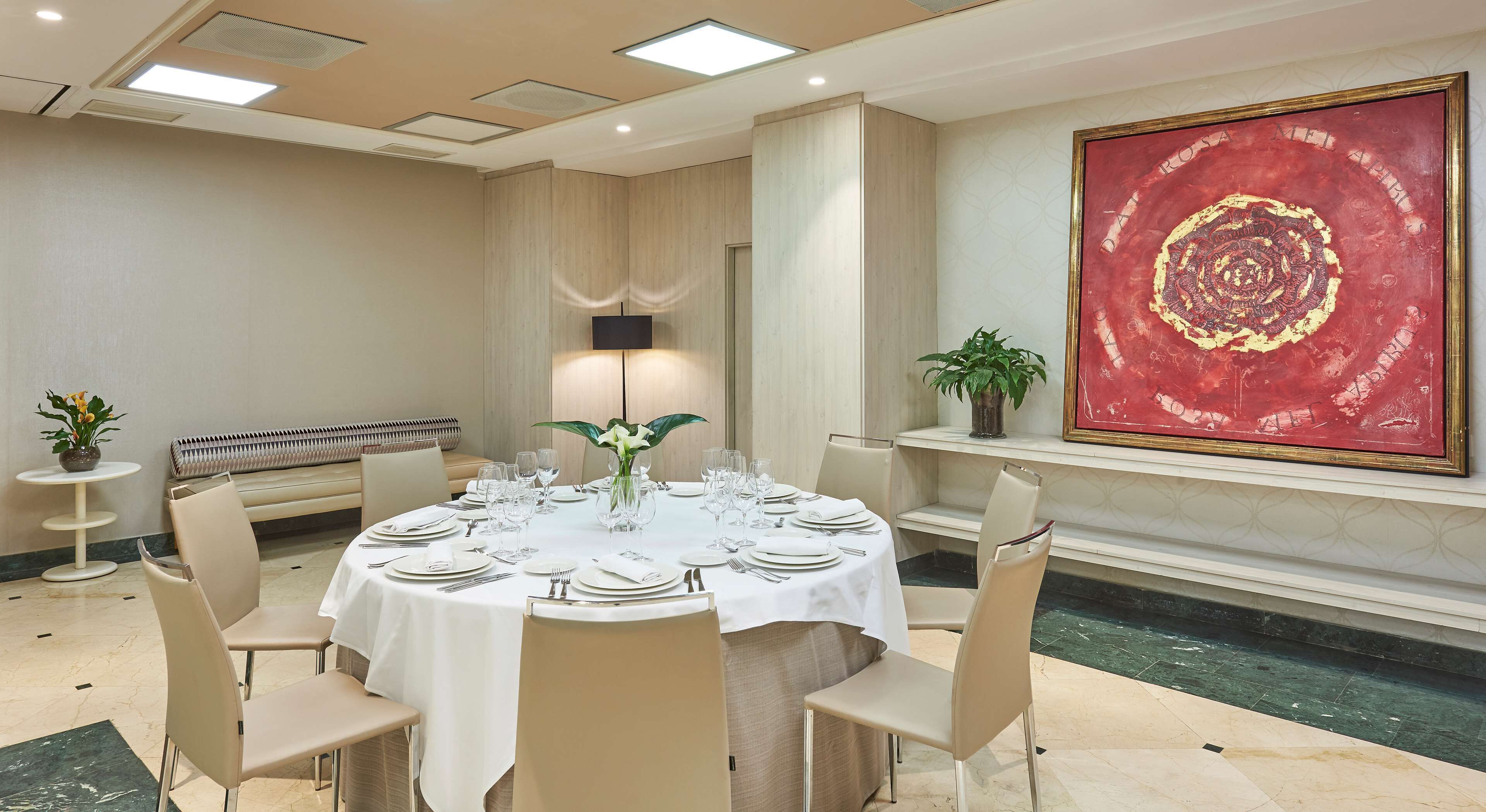 A conference room with a round table set for a meeting, a large red painting on the wall, and a potted plant on a shelf.