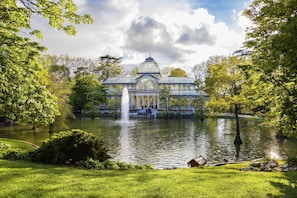 A large glass conservatory with a fountain in the foreground, surrounded by lush greenery and a pond.