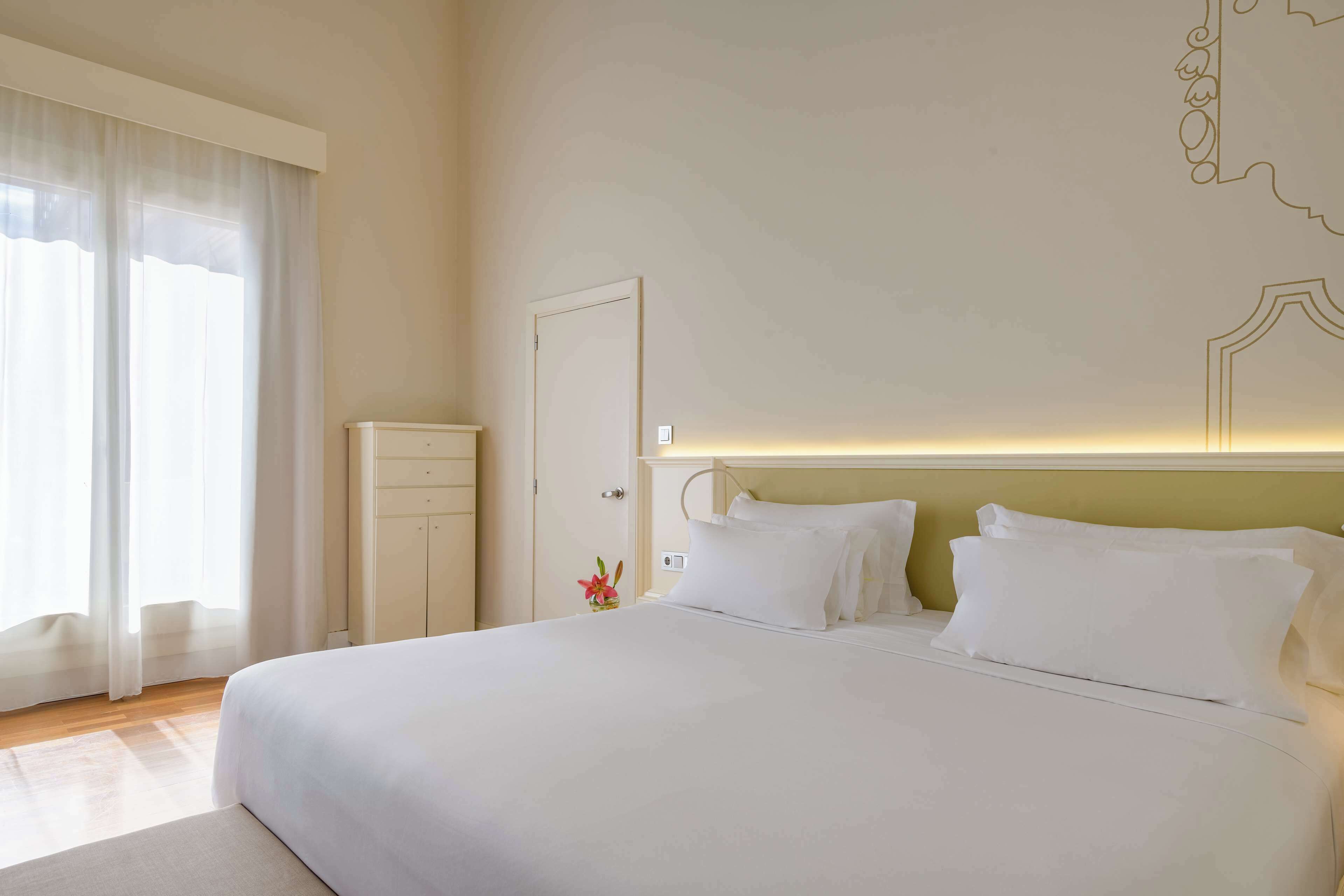 A large bed with white linens, a bedside table, and a nightstand with a vase of flowers.