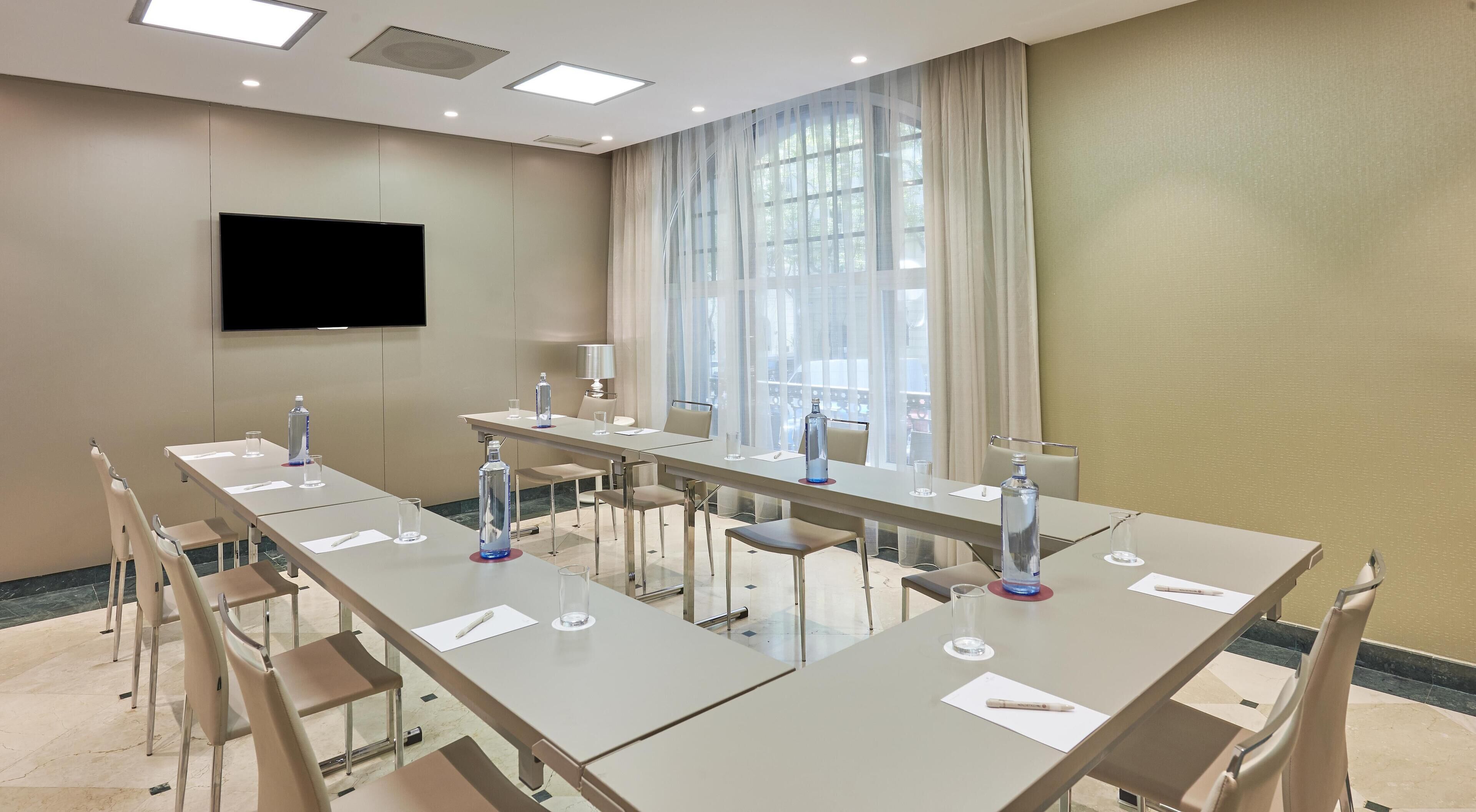 A conference room with a long table, chairs, a flat-screen TV, and large windows with sheer curtains.