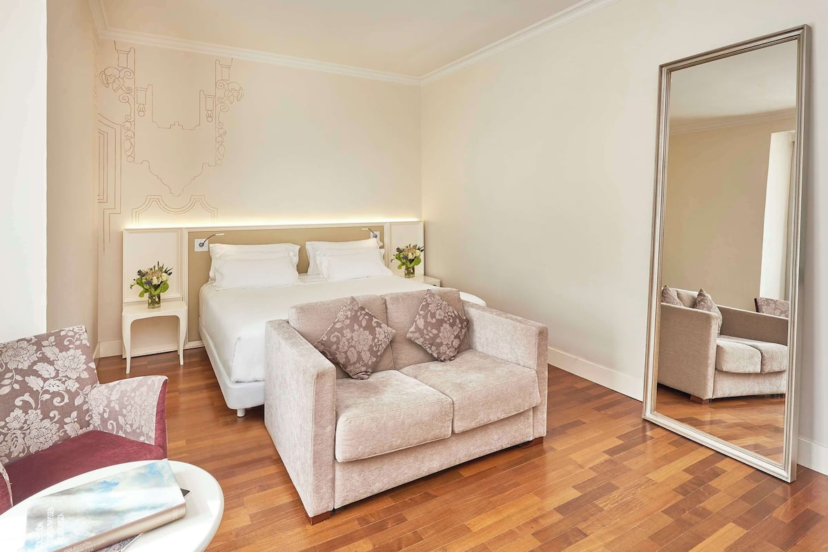 Una habitación de hotel con cama, sofá, sillón, espejo y piso de madera.