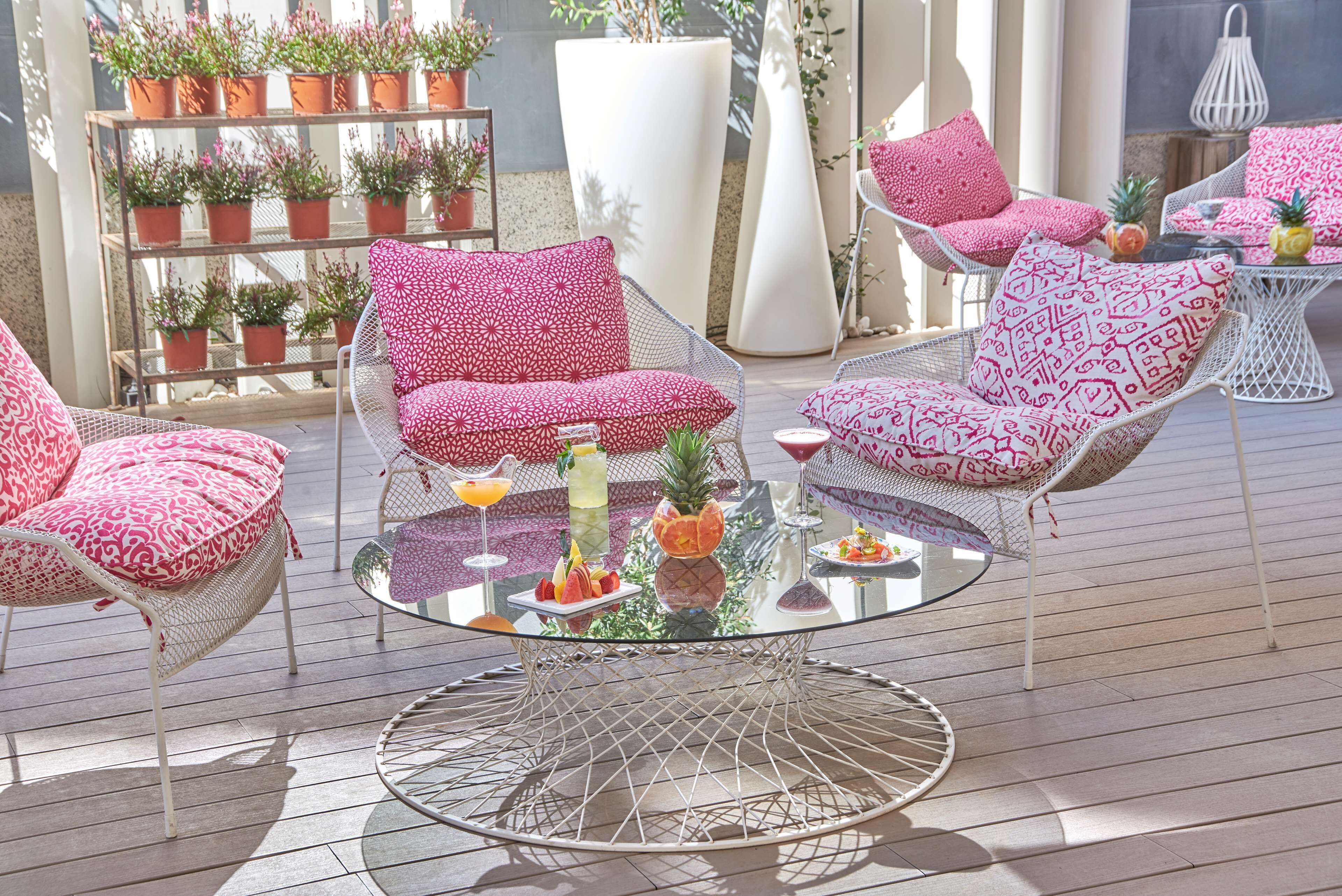 A sunlit outdoor seating area with wicker chairs and a glass table, surrounded by potted plants and a white column.