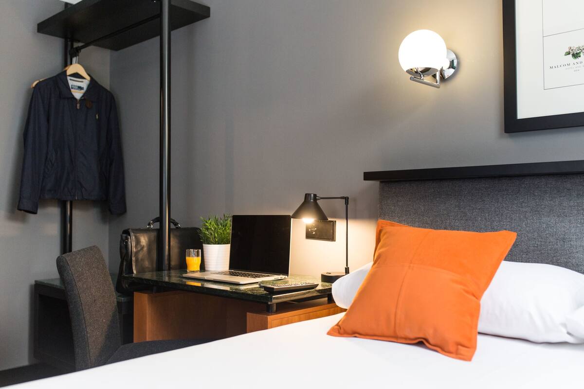 Habitación de hotel ordenada con cama, escritorio con una computadora portátil, silla, abrigo colgado y lámpara de pared.