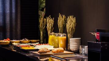 A buffet with various food items, including bread, vegetables, and a fruit salad. There are also several glasses of juice and a coffee machine.