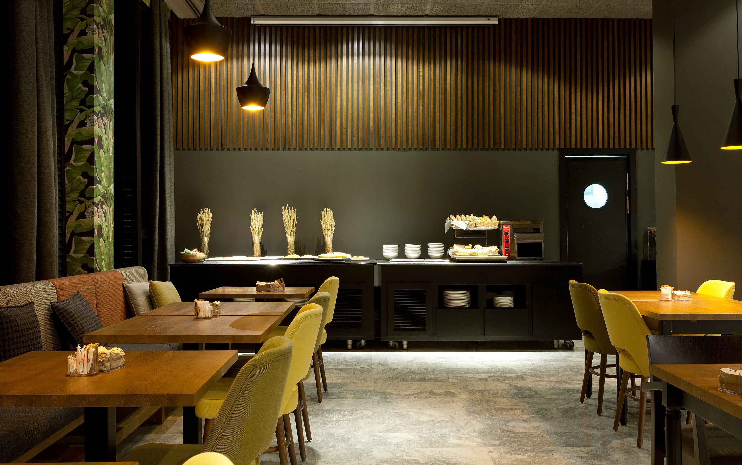 A restaurant with wooden tables and yellow chairs, a counter with food and a coffee machine, and a wall with vertical slats.