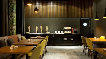 A restaurant with wooden tables and yellow chairs, a counter with food and a coffee machine, and a wall with vertical slats.