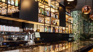 Zona de bar con estantes llenos de botellas de licores, una máquina de café y una barra con una pantalla de menú.