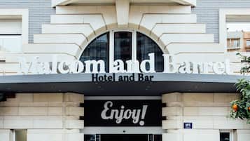 Een hotelentree met een bord waarop 'Malcolm and Barret Hotel and Bar' staat en een 'Enjoy!' bord boven de deur.