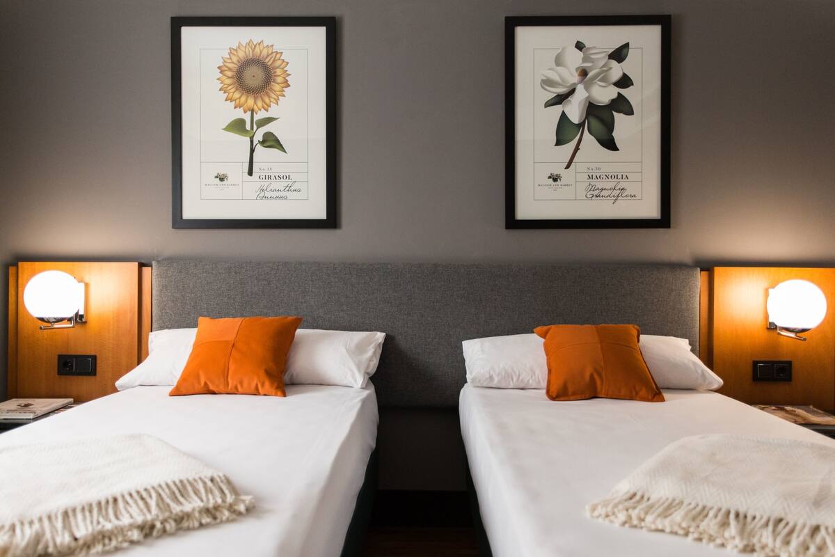 Habitación de hotel con dos camas, cada una con ropa de cama blanca y almohadas naranjas, e impresiones botánicas enmarcadas encima.