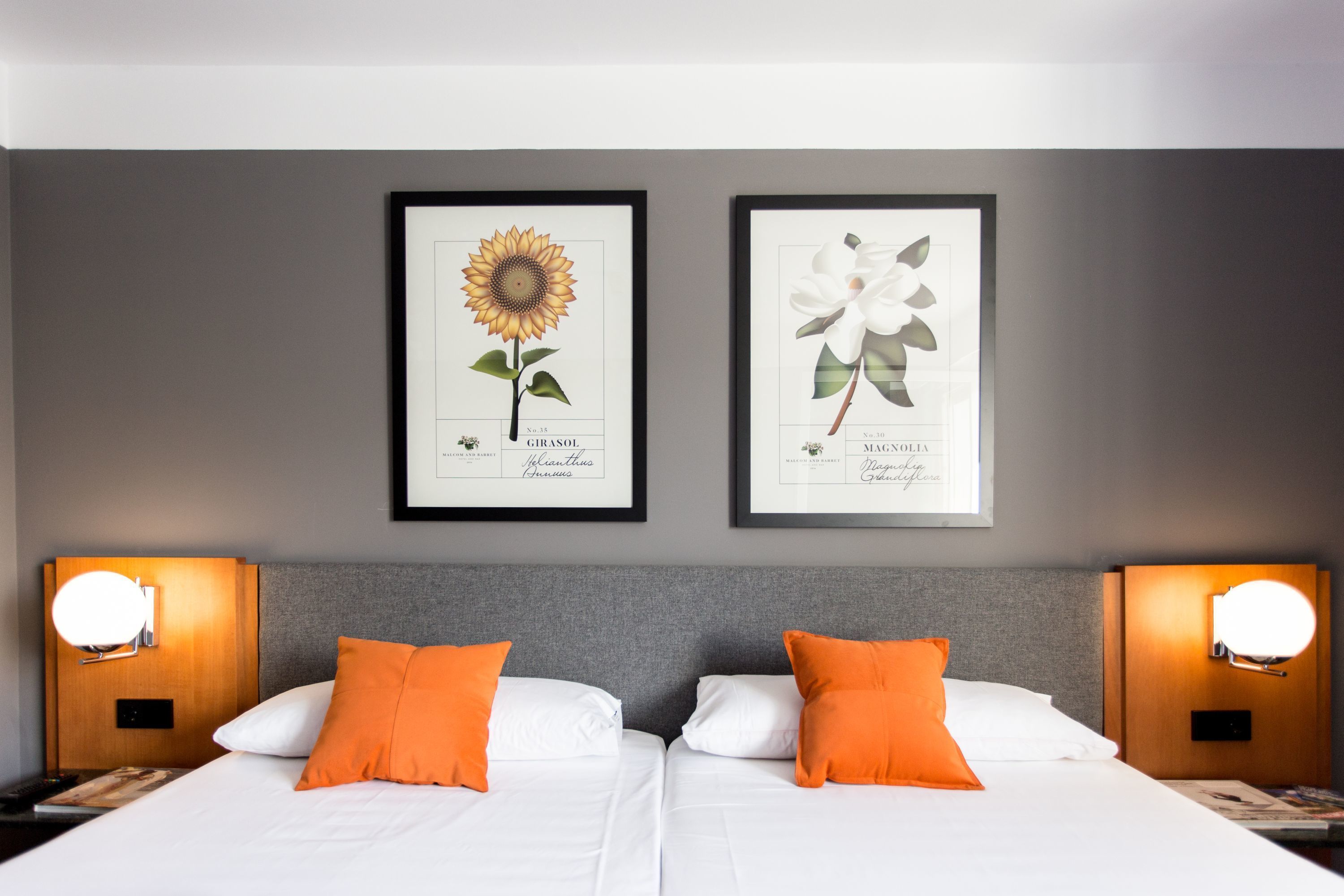 Une chambre d’hôtel avec deux lits, chacun agrémenté de draps blancs et d’oreillers orange. Des reproductions encadrées de motifs botaniques ornent le mur au-dessus des lits.