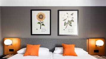 Une chambre d’hôtel avec deux lits, chacun agrémenté de draps blancs et d’oreillers orange. Des reproductions encadrées de motifs botaniques ornent le mur au-dessus des lits.