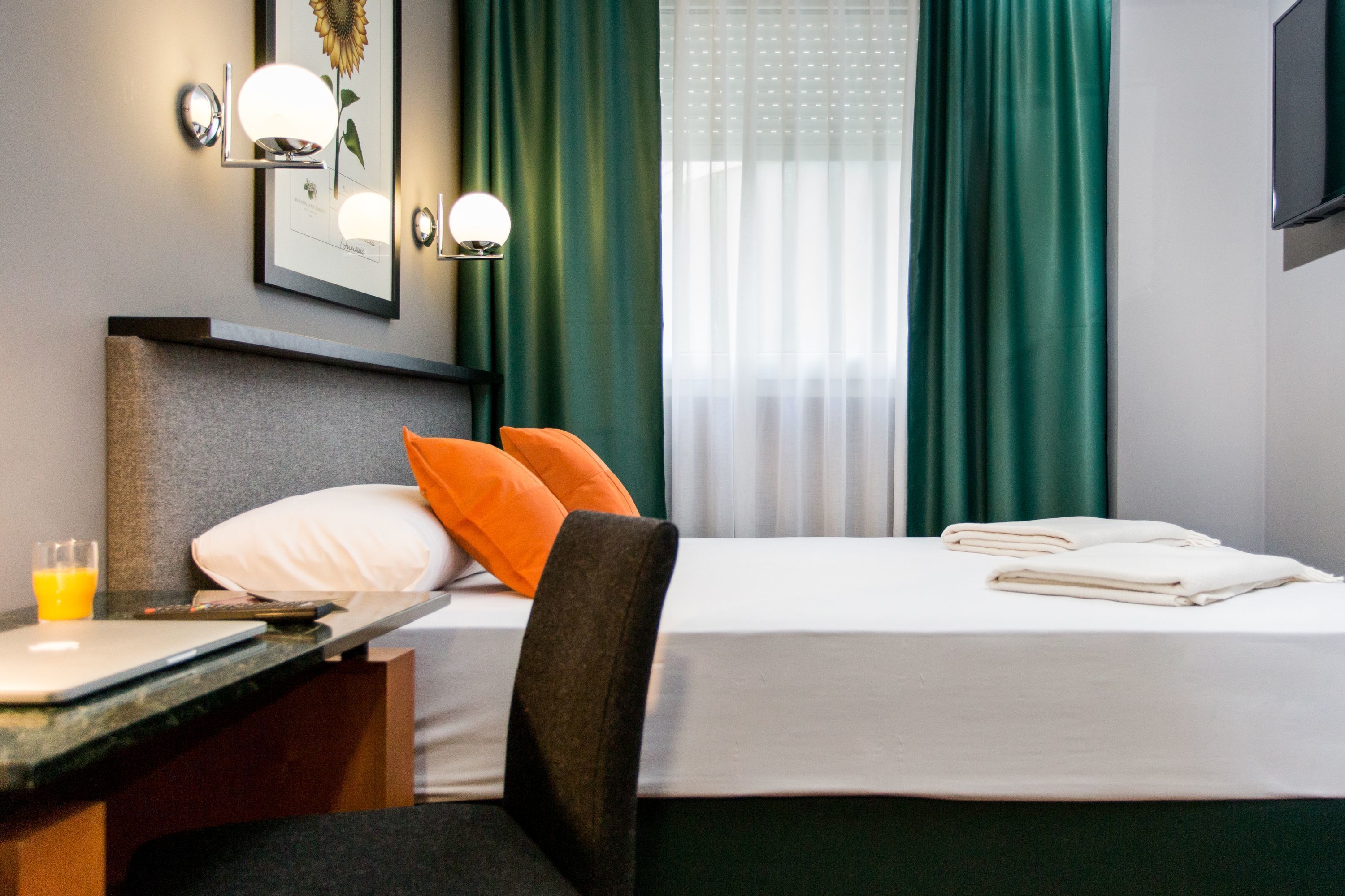 A hotel room with a bed, a desk with a laptop, a chair, a framed picture on the wall, and a window with curtains.