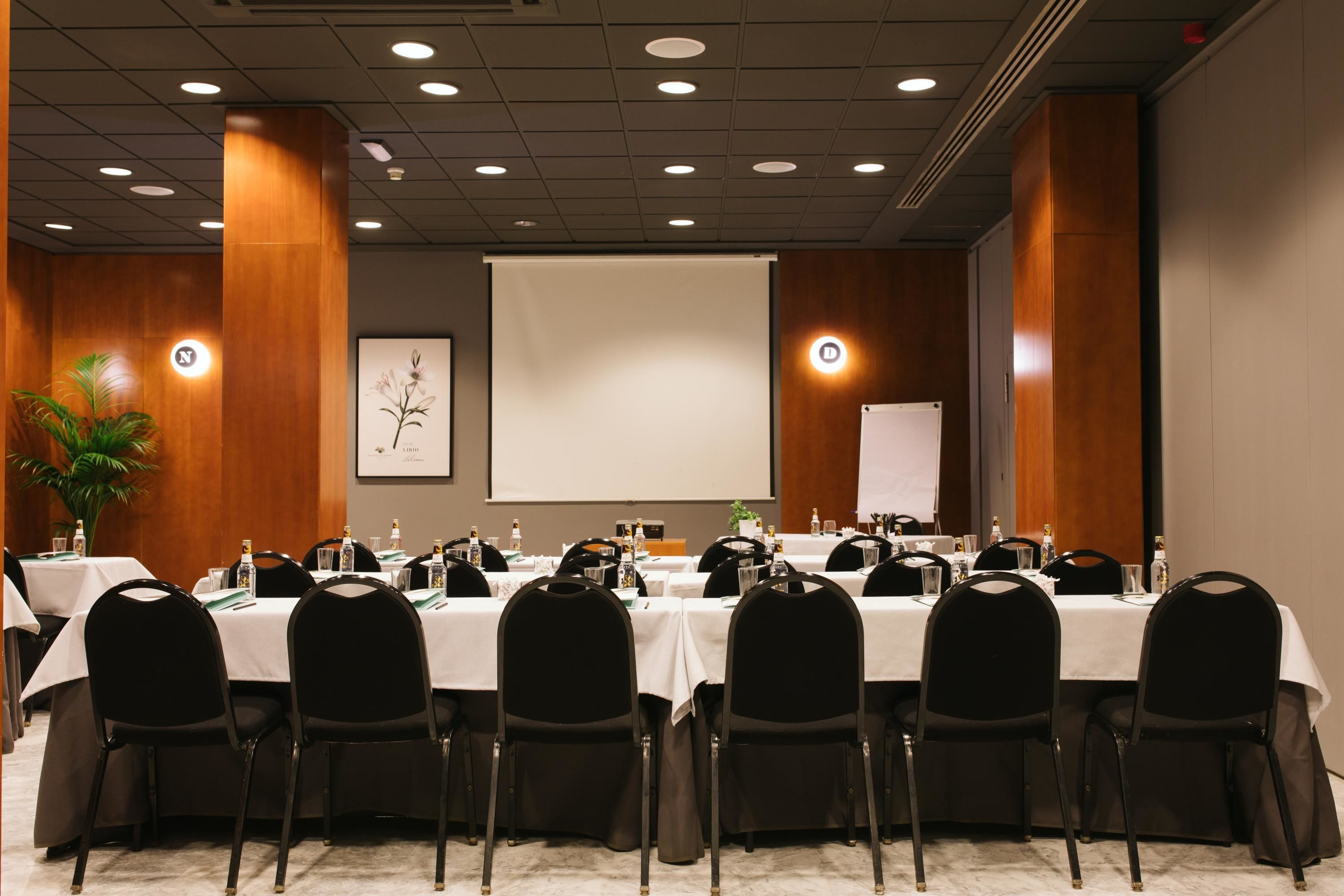 Un salón de conferencias con varias mesas, sillas negras y una pantalla para proyector.