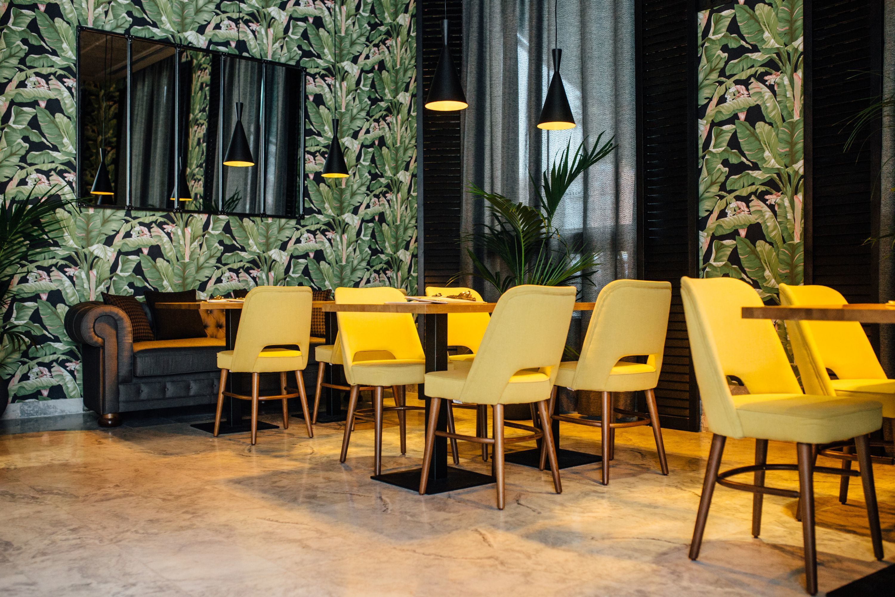Een eetgedeelte met gele stoelen en een bank, een grote spiegel, en een muur met een bladerenpatroon.