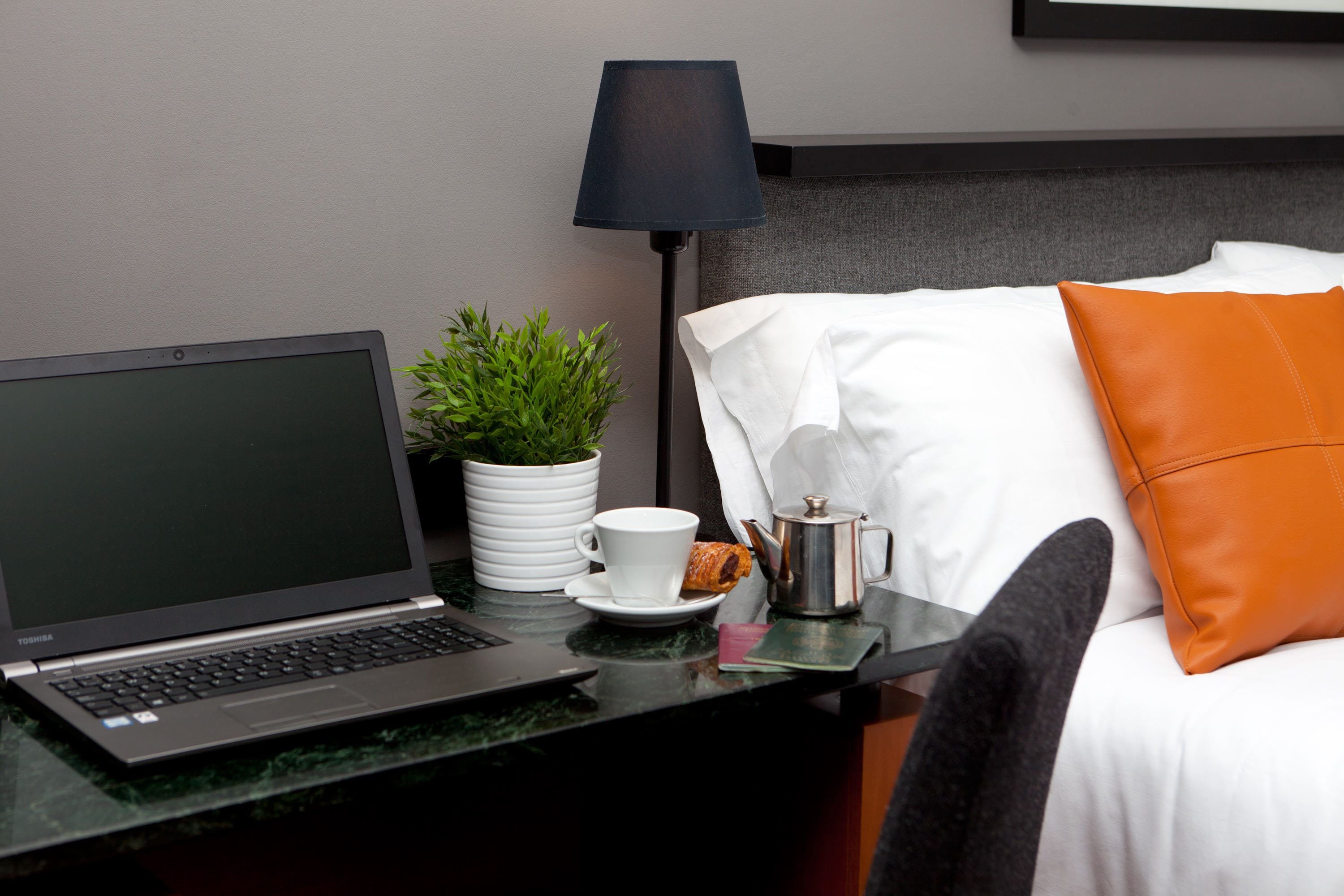 Una habitación de hotel con cama, escritorio, computadora portátil, taza de café, planta y lámpara.