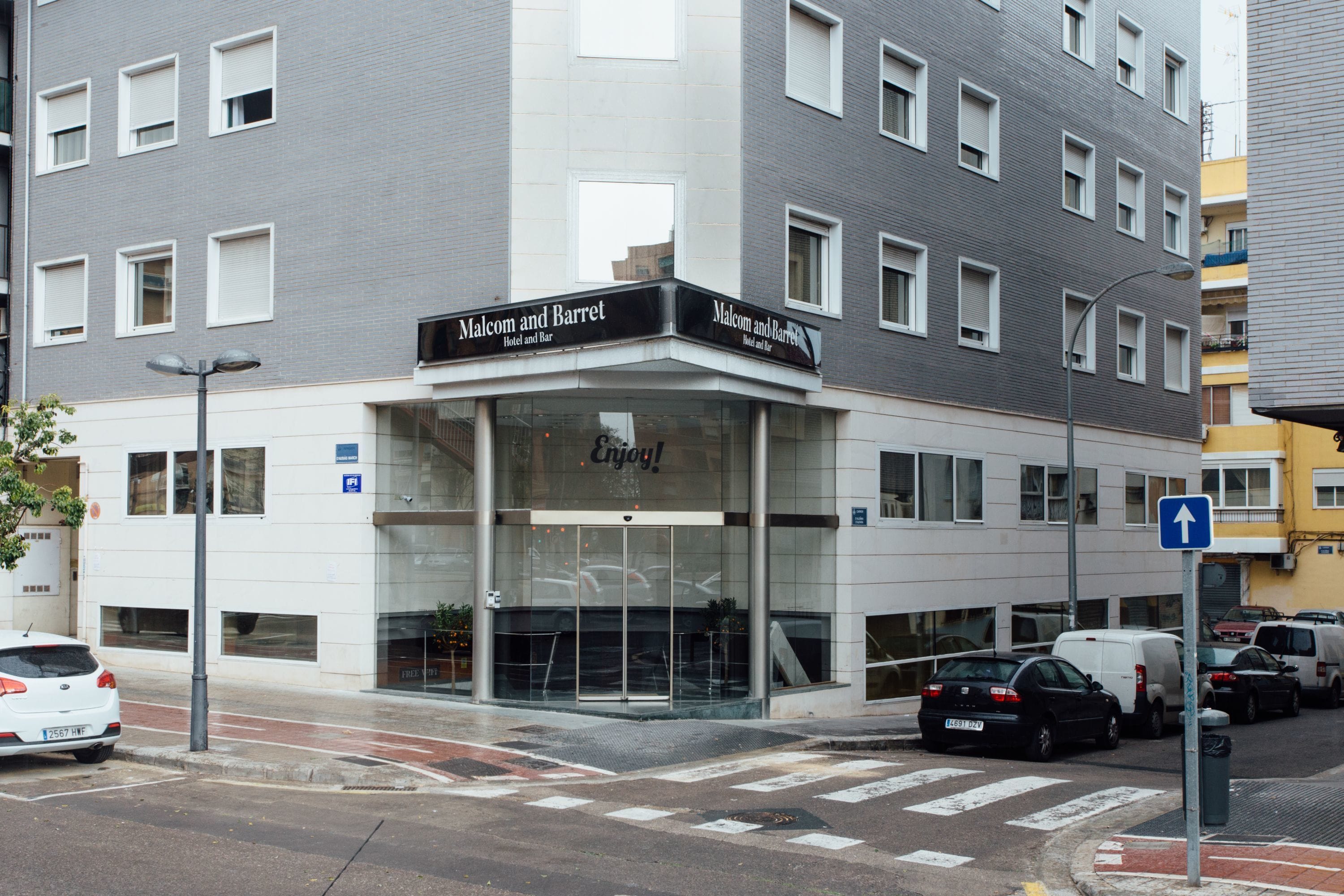A hotel named 'Malcom and Barret' with a glass entrance, parked cars, and a pedestrian crossing.